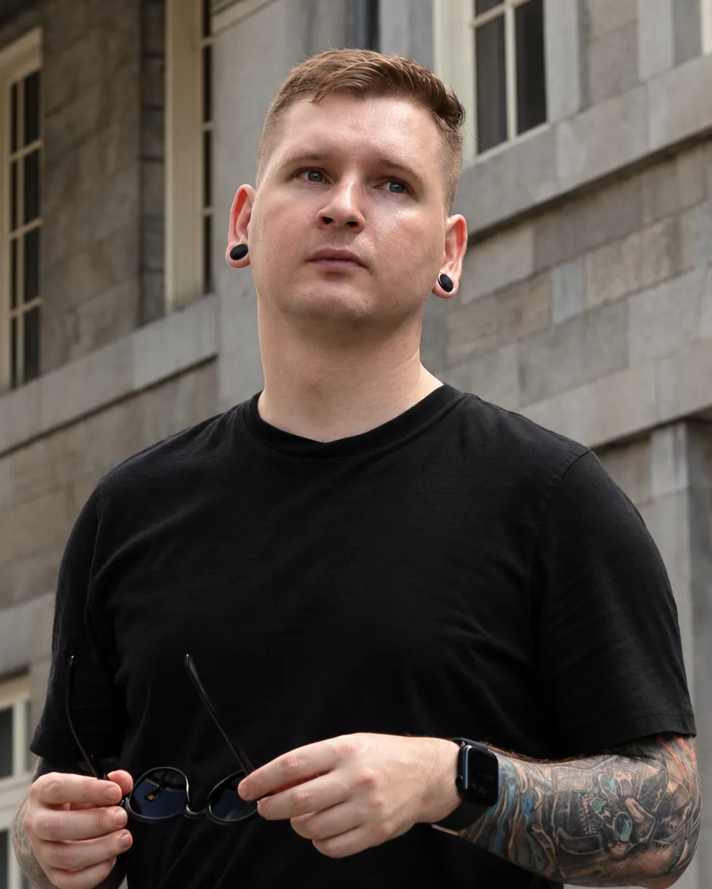 Ross, standing outside in a black t-shirt with a tattooed forearm